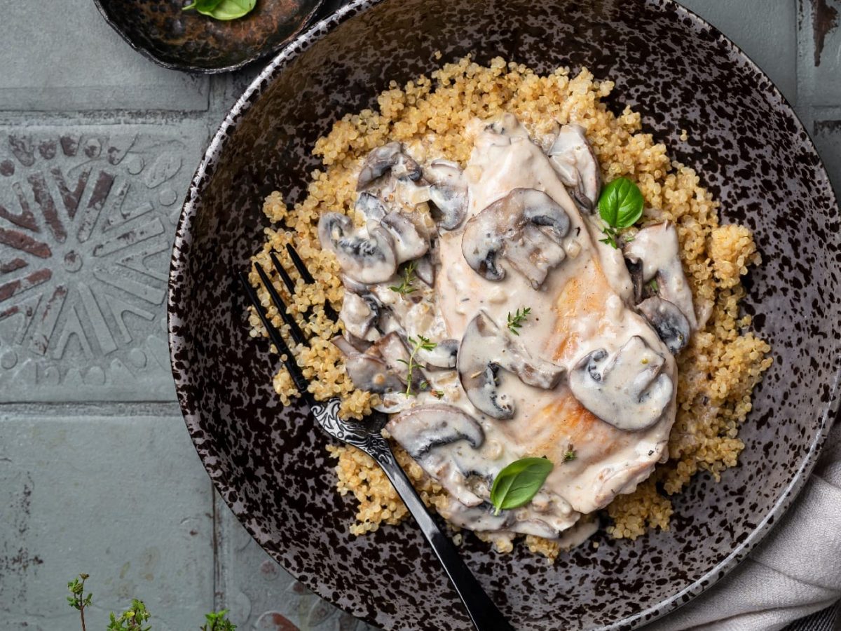 Ein schwarzer Teller mit Quinoa und dem Pilz-Rahm auf grauen Fliesen mit einer schwarzen Gabel.