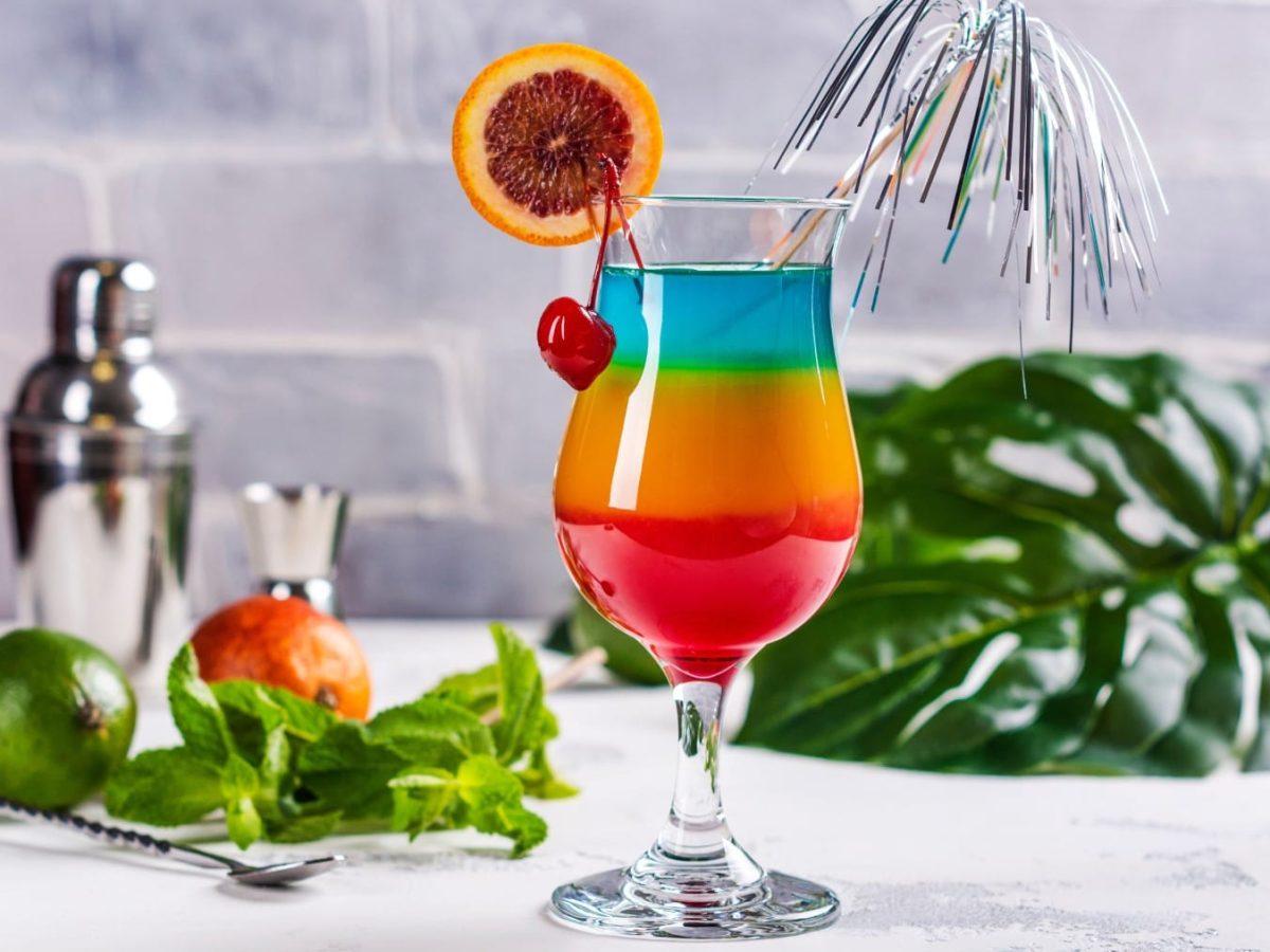 Ein Glas mit Regenbogen-Cocktail (unten rot, Mitte gelb-orange, oben blau), garniert mit Kirsche und Blutorangenscheibe vor hellem Hintergrund.