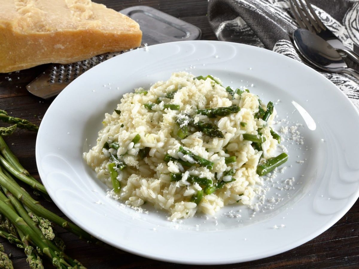 Draufsicht: ein Teller Risotto mit Wildspargel auf schwarzem Untergrund. Daneben liegen Wildspargel-Stangen, Parmesan und Besteck.