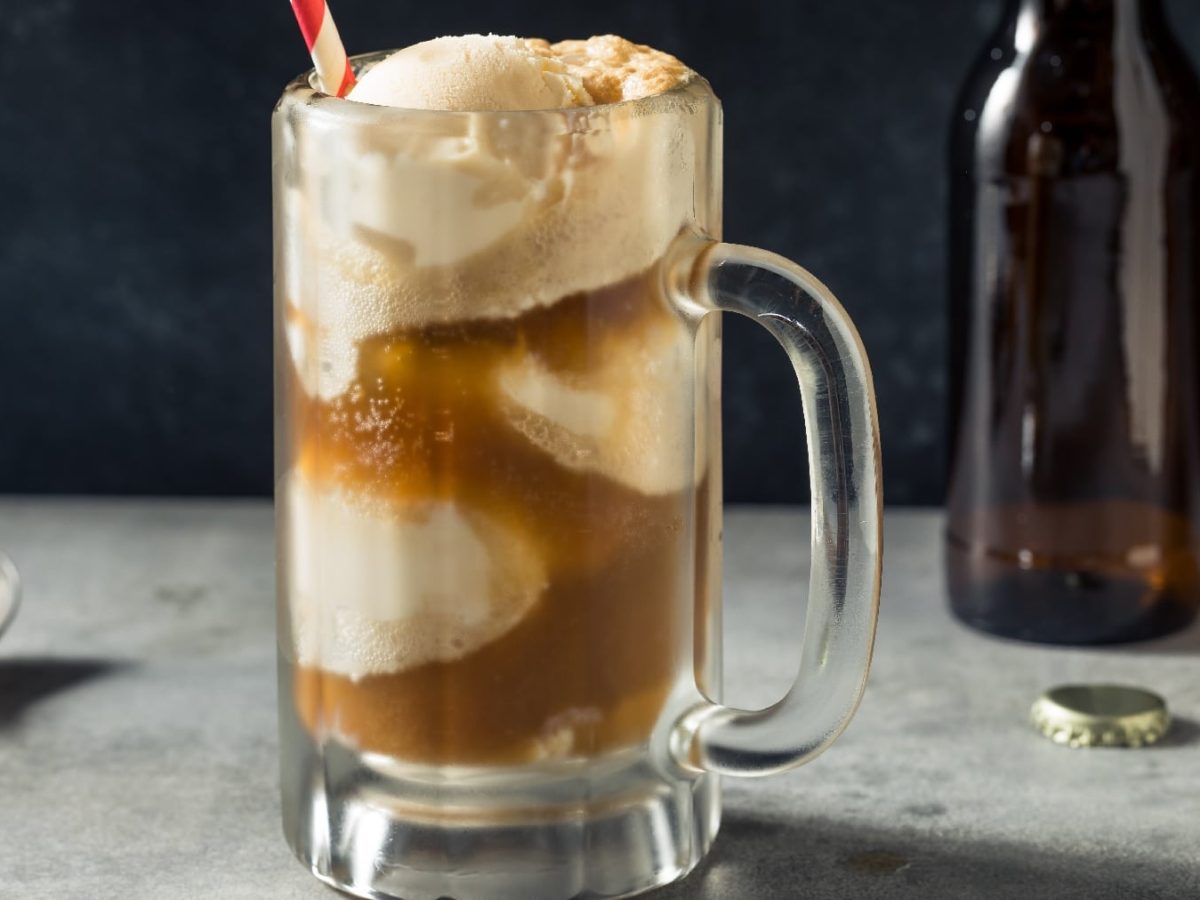 Ein Glas mit Root-Beer-Float auf einem grauen Untergrund mit Bierflasche und schwarzem Löffel.