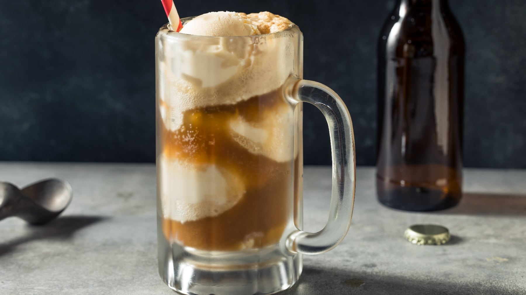 Ein Glas mit Root-Beer-Float auf einem grauen Untergrund mit Bierflasche und schwarzem Löffel.