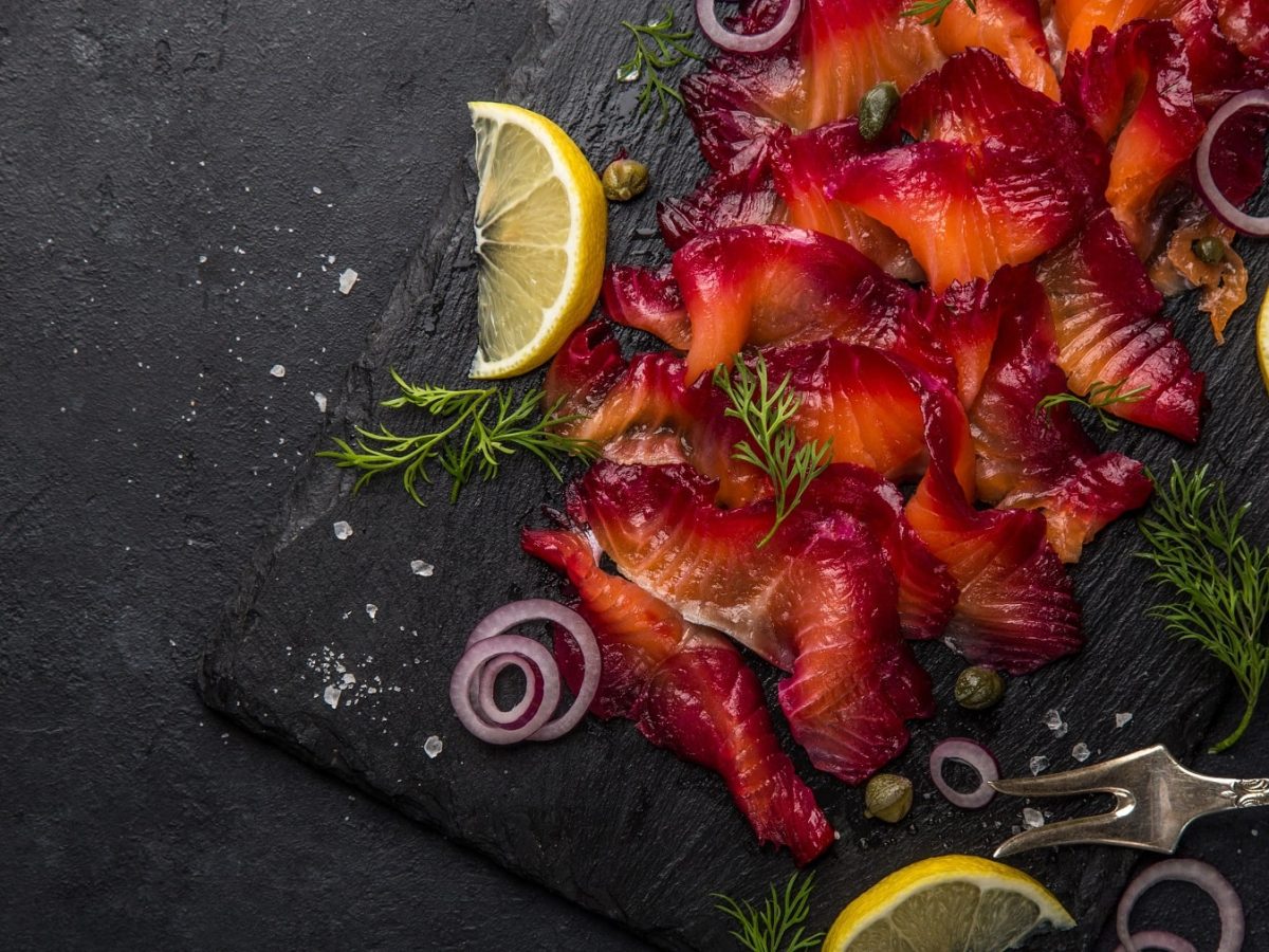 Der in Rote-Bete gebeizte Lachs aufgeschnitten auf einer schwarzen Schieferplatte mit Kapern und Zitronenscheiben.