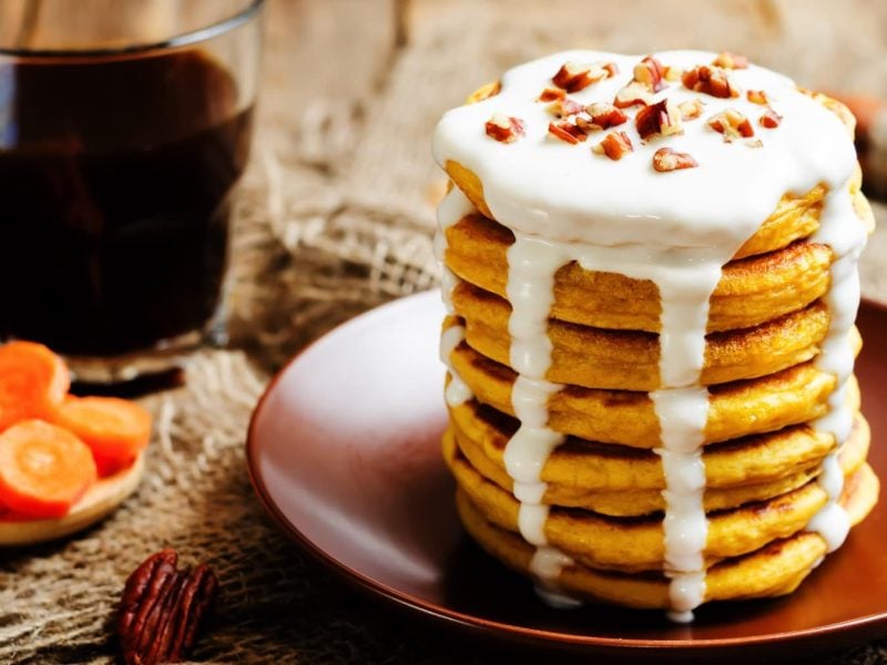 Ein Stapel Rübli-Pancakes mit Frosting-Sauce und gehackten Nüssen auf einem braunem Teller neben einem Glas Kaffee und Möhrenscheiben.