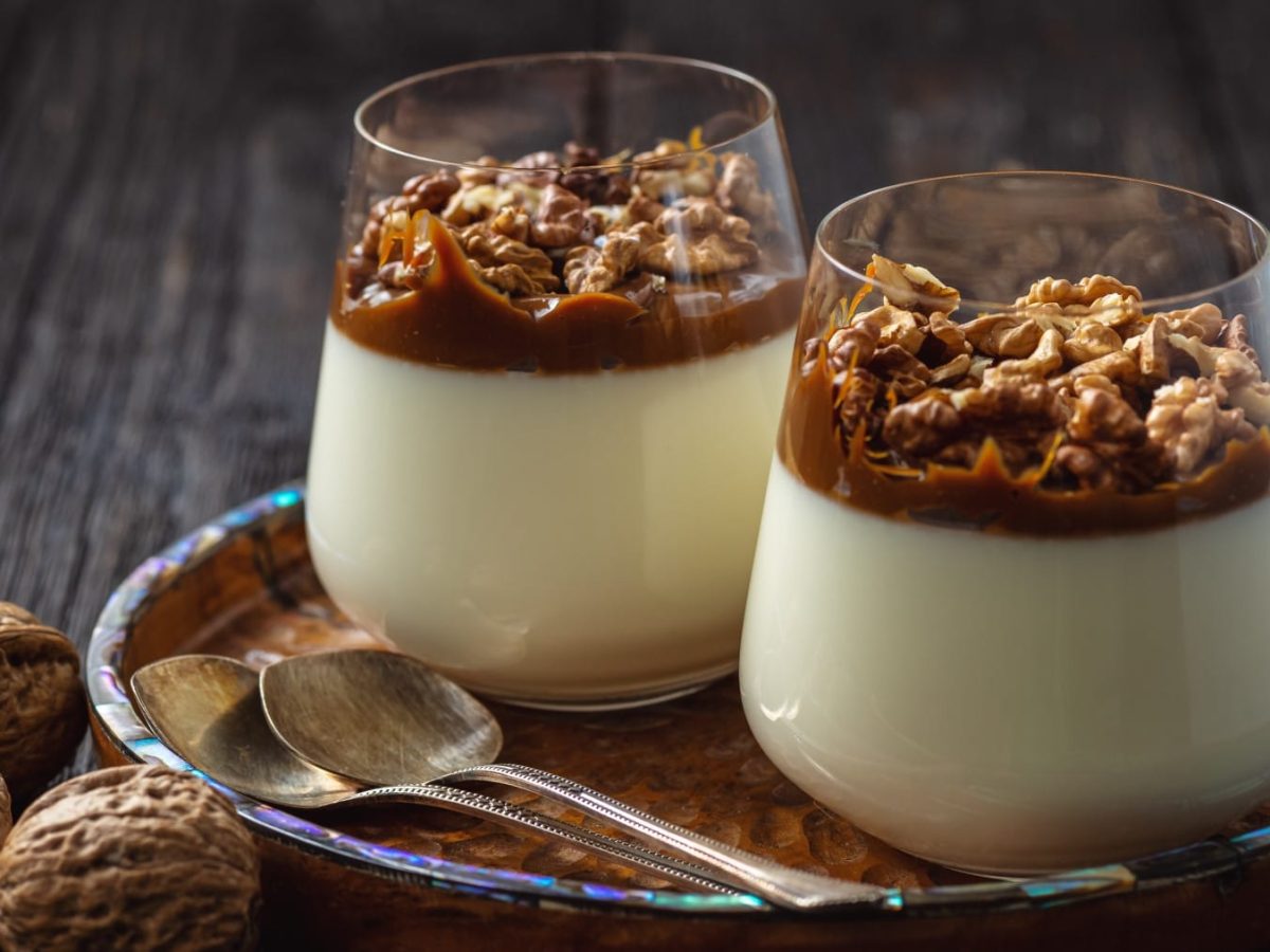 Zwei Gläser Sahnepudding mit Dulce de Leche und Walnüssen auf einem Tablett, daneben Löffel und Walnüsse vor Holzhintergrund.