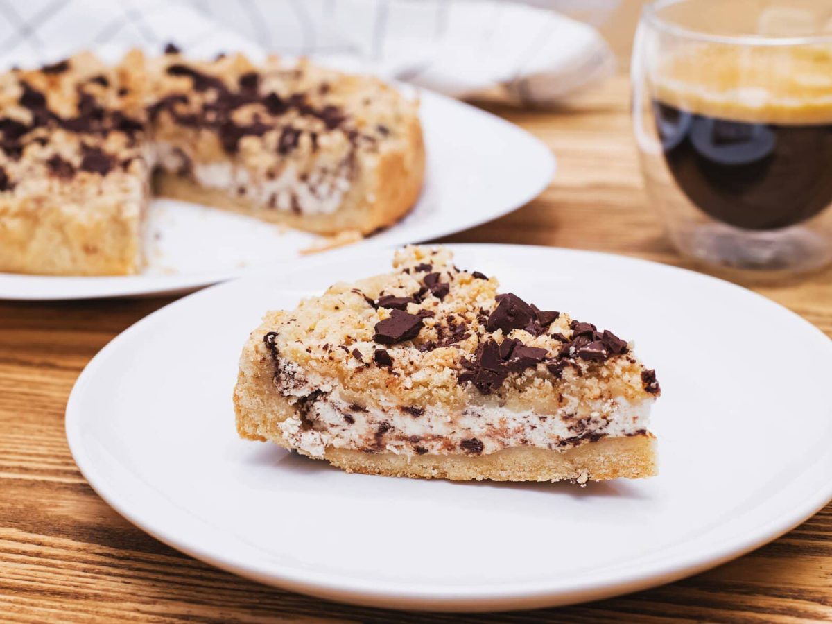 Ein Stück Sbriciolata alla Ricotta auf weißem Teller auf Holztisch. Im Hintergrund restlicher Kuchen und eine Tasse Espresso. Draufsicht.