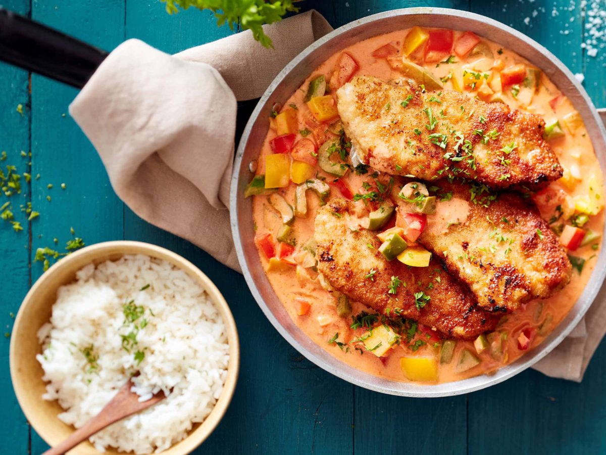 Schweineschnitzel auf Paprika-Rahm in einer Pfanne, daneben eine Schüssel mit Reis vor blauem Hintergrund in der Draufsicht.