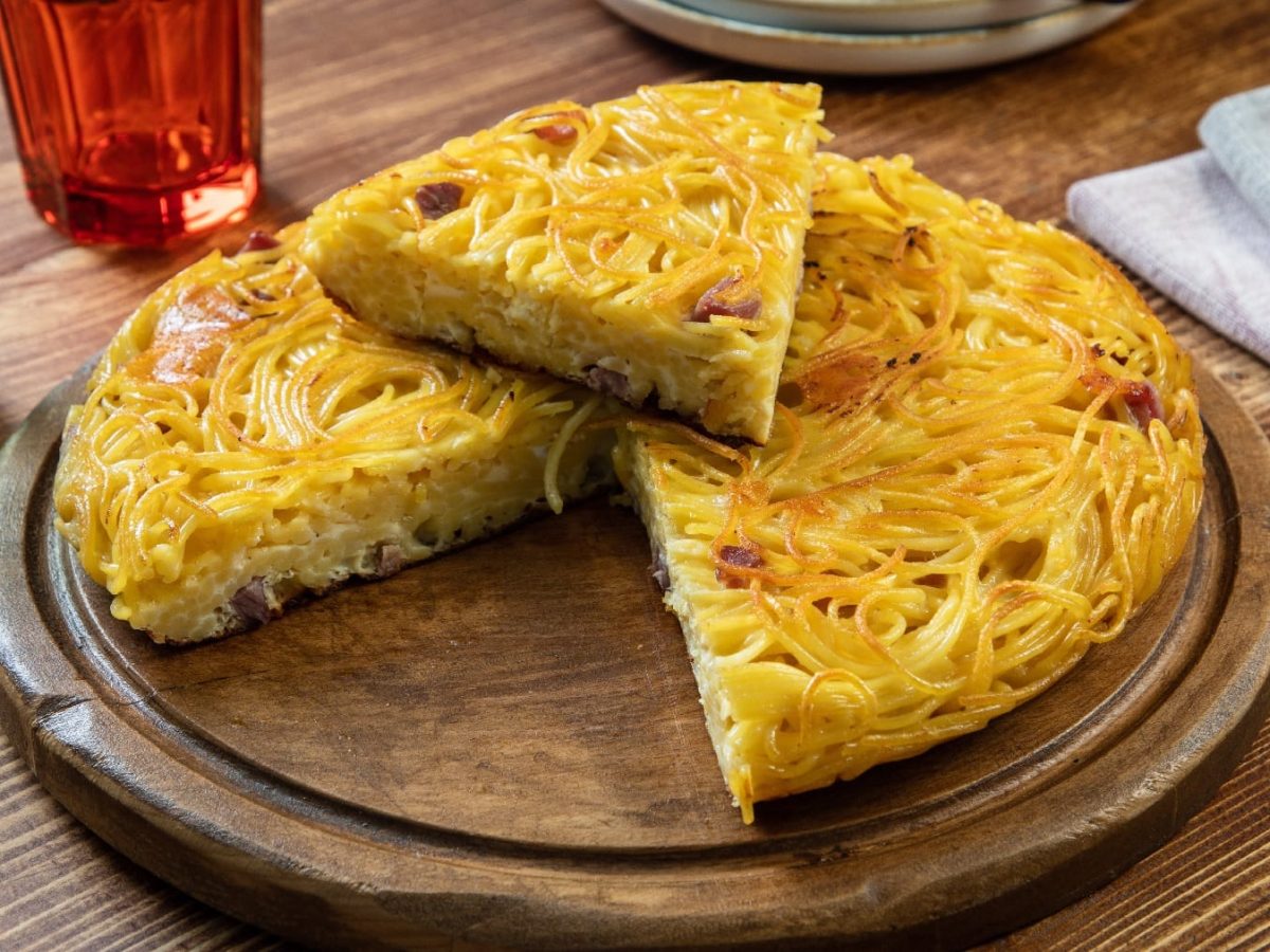 Der Spaghetti-Carbonara-Kuchen auf einem Holzbrett und einem Holztisch. Im Hintergrund unscharf zwei Gläser.