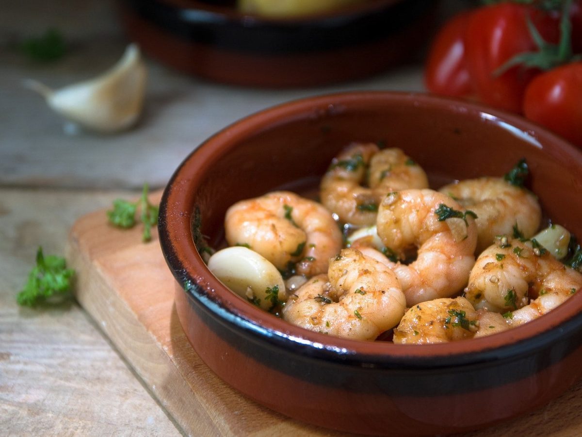 Frontalsicht: Gebratene Shrimps mit Knoblauch und Petersilie in Öl in einer Tonschüssel. Im Hintergrund sind rote Chilis und Knoblauchzehen.