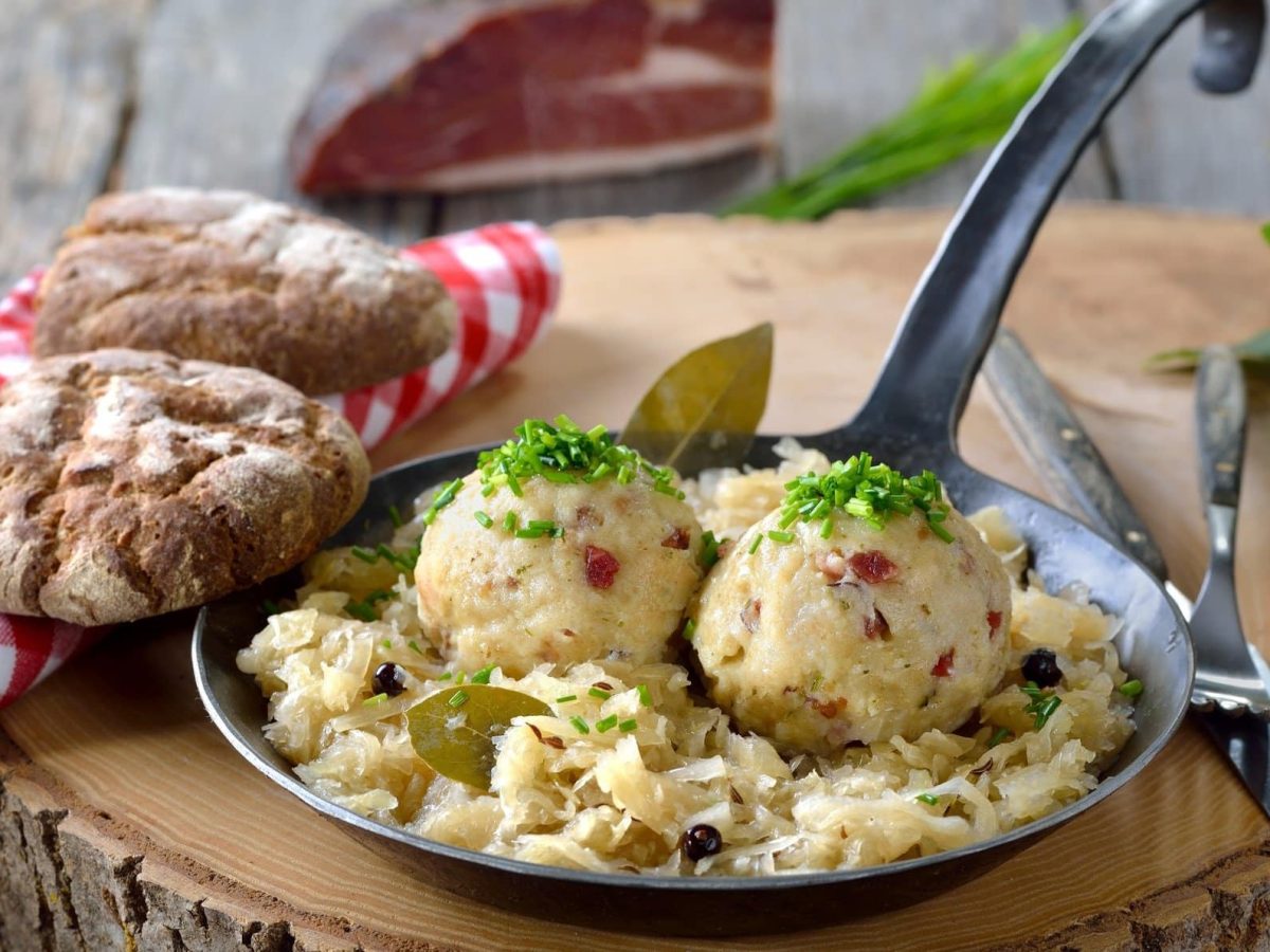 Frontalsicht: Auf einem Holztisch steht eine gusseiserne Pfanne Speckknödel mit Sauerkraut. Drum herum liegen Gewürze und ein weiß-rot kariertes Tuch.