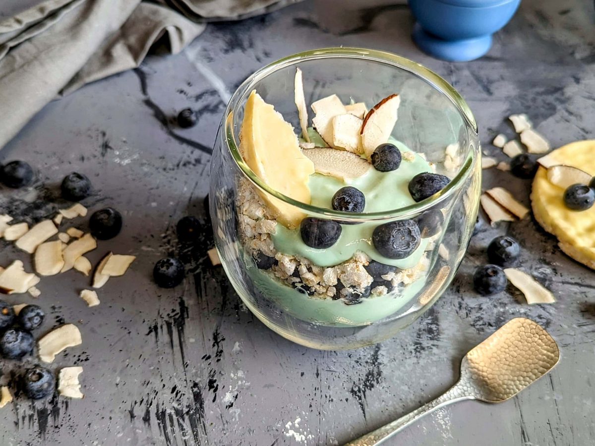 Ein Glas mit dem geschichteten Frühstück aus Spirulina-Joghurt, Blaubeeren , Resiwaffel und Kokoschips auf einem grauen Marmoruntergrund.