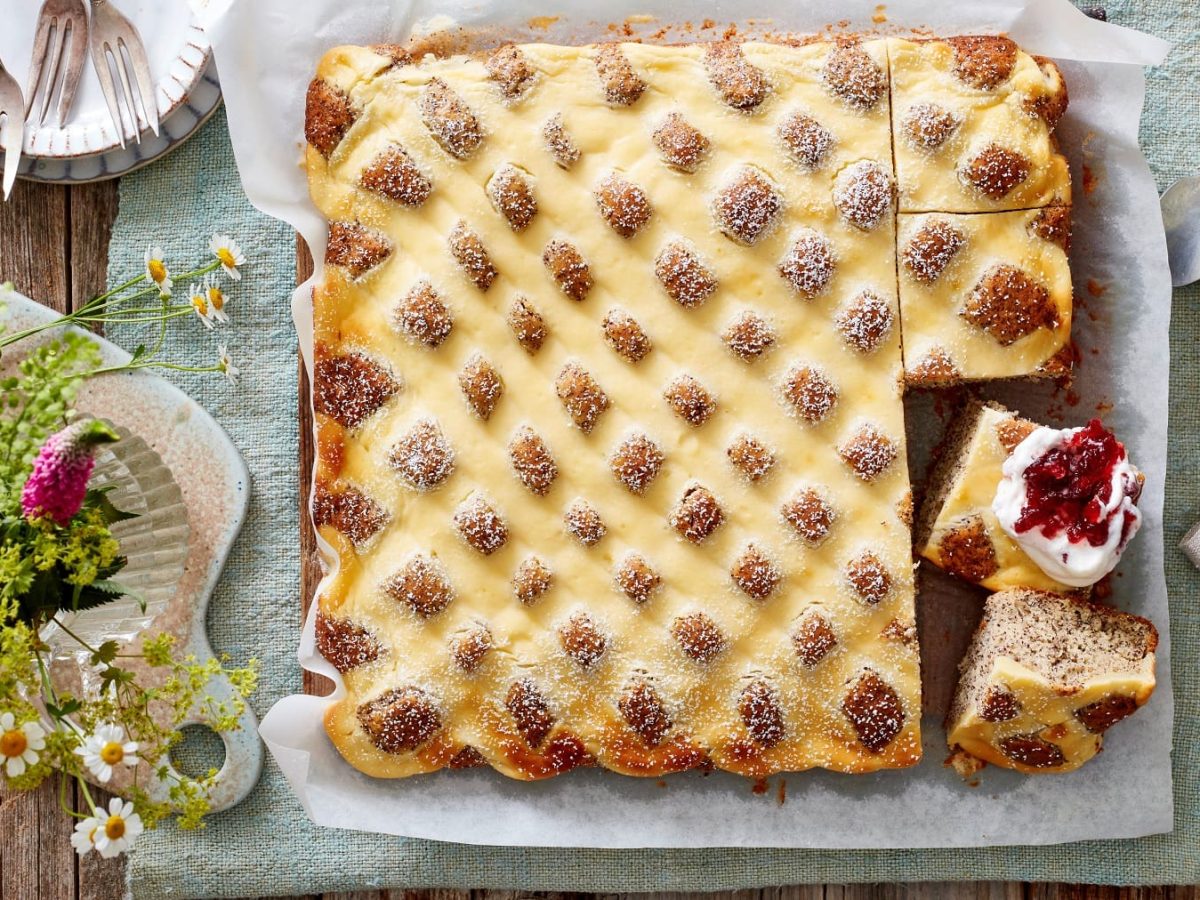 Angeschnittener Steppdecken-Mohnkuchen auf Backpapier vor hellem Hintergrund in der Draufsicht, daneben Blumen, Geschirr und Quark mit Beeren.