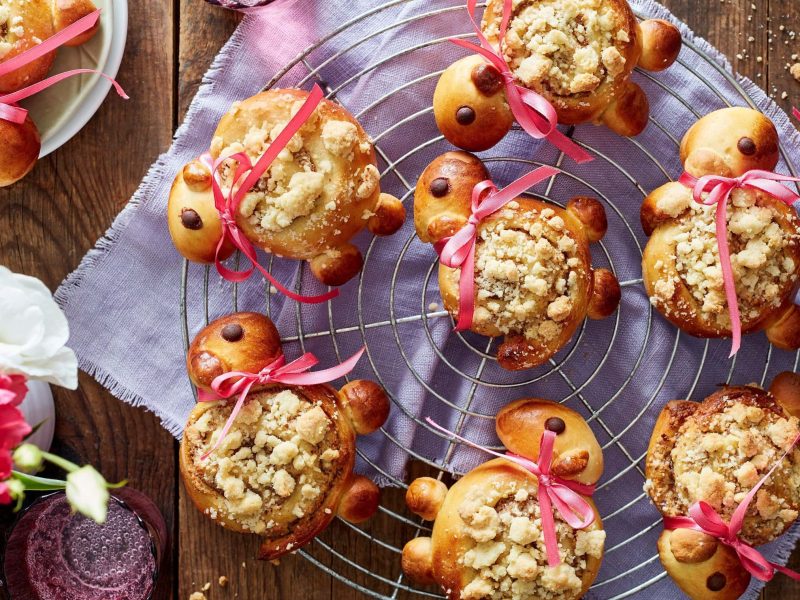 Mehrere Streusel-Lämmer mit rosa Schleifen auf einem Gitter vor lila Küchentuch in der Draufsicht.