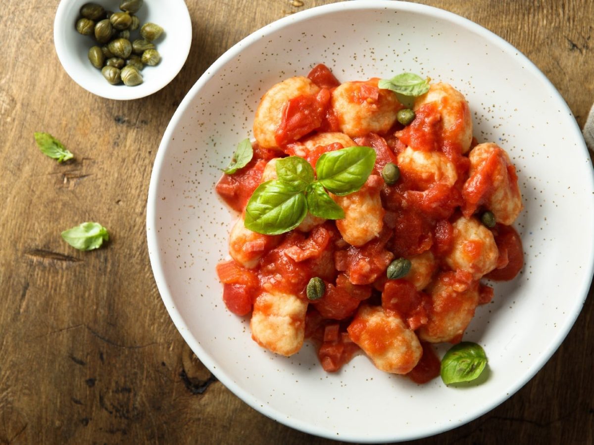 Ein weißer Teller mit den Mehl-Gnocchi und der Sucuk-Tomatensauce mit Kapern auf einem Holztisch.