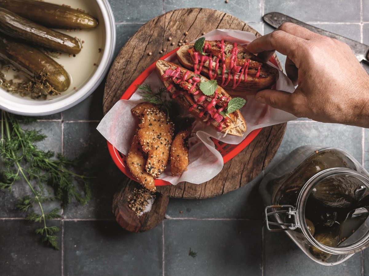 Sum Sum Schnitzelim, kleine Schnitzel in geröstetem Zatar-Pita-Brot, nach denen eine Hand greift. Daneben eine Schüssel und ein Glas Gewürzgurken. Alles Draufsicht.