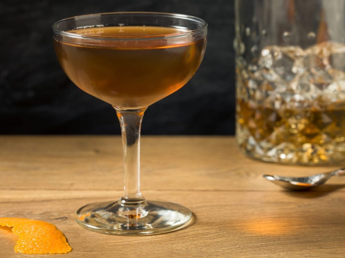 Tipperary Cocktail No.1 mit Proper No. Twelve im Glas auf Holztisch. Im Bild ein Whiskey Glas, Orange und Orangenzeste.