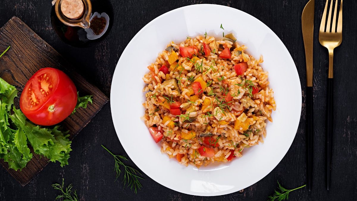 Ein weißer Teller mit dem Tomatenreis auf einem dunklen Untergrund. Daneben goldenes Besteck und ein Holzbrett mit einer Tomate und etwas Salat.