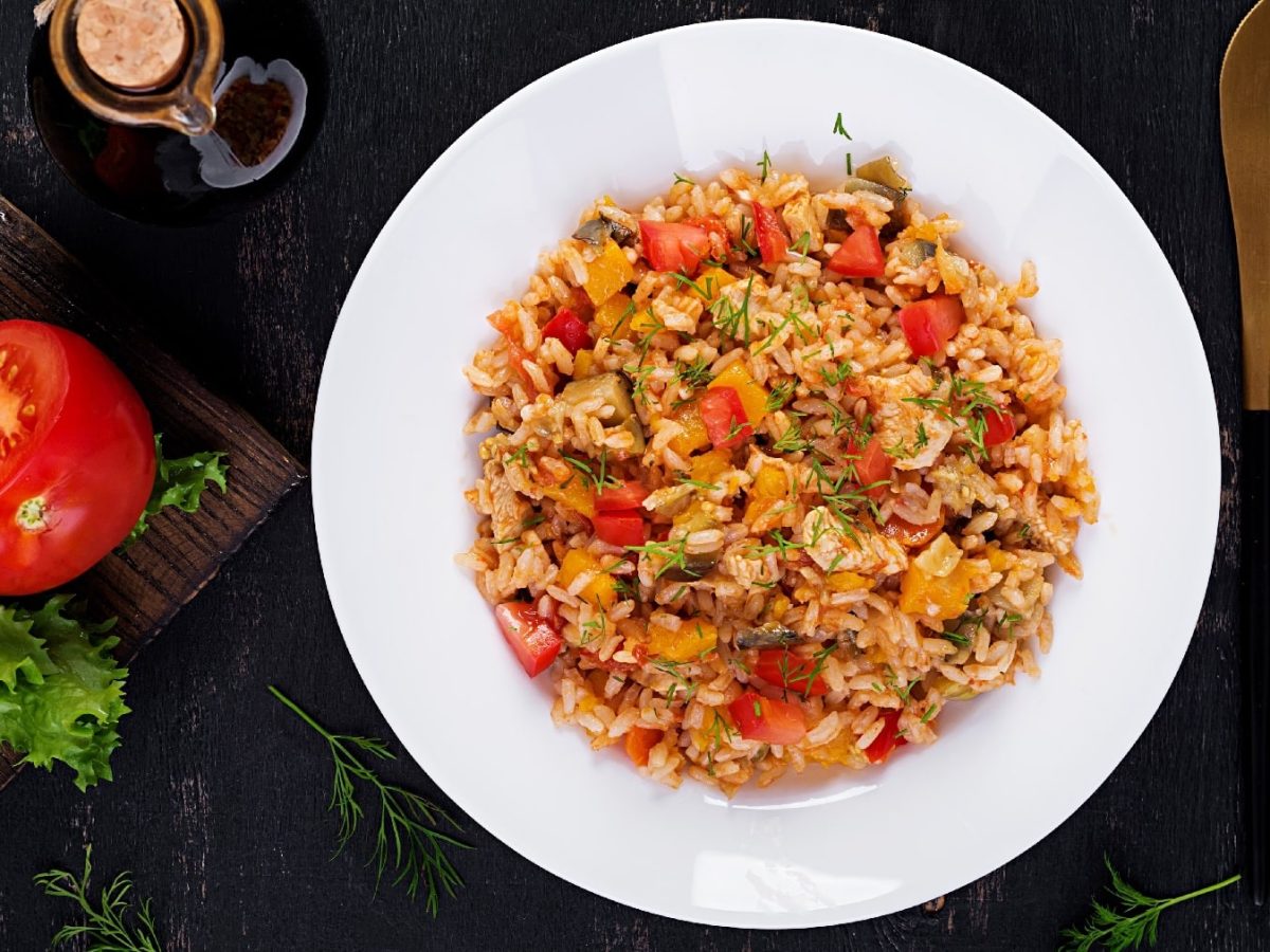 Ein weißer Teller mit dem Tomatenreis auf einem dunklen Untergrund. Daneben goldenes Besteck und ein Holzbrett mit einer Tomate und etwas Salat.