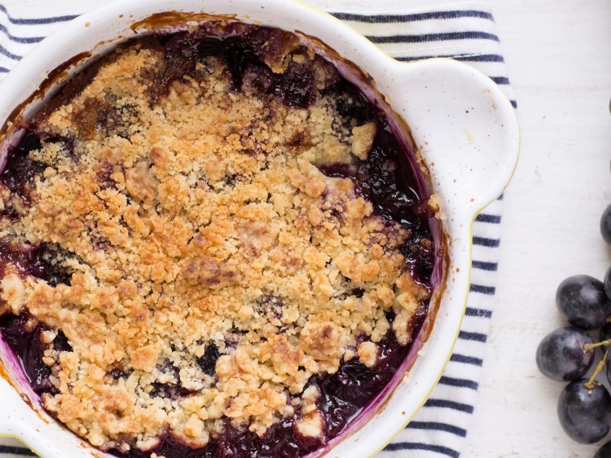 Eine weiße Auflaufform mit dem Trauben-Crumble auf einer blau-weiß-gestreiften Tischdecke.