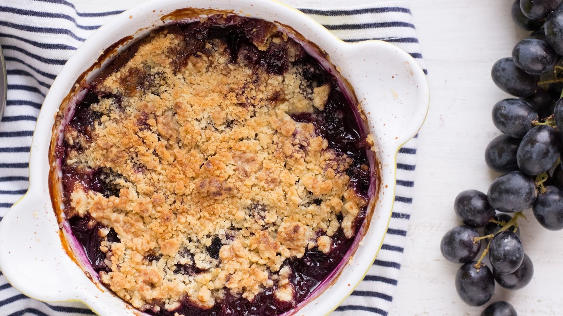 Eine weiße Auflaufform mit dem Trauben-Crumble auf einer blau-weiß-gestreiften Tischdecke.