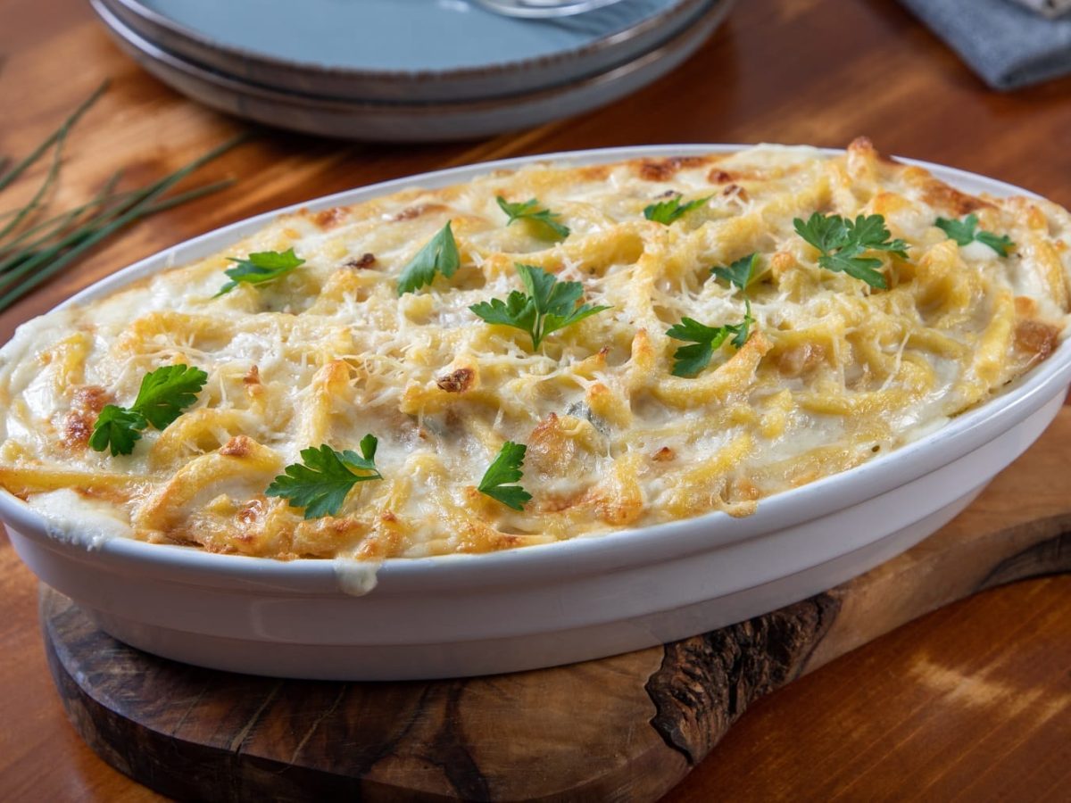 Eine weiße Auflaufform mit den überbackenen Spätzle auf einem Holzbrett und einem Holztisch mit zwei Tellern, Gabeln und einem Bund Schnittlauch.