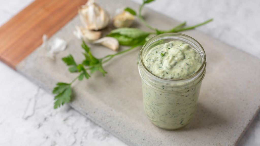 Ein Glas vegane Frankfurter Grüne Soße auf einem Schneidebrett, dahinter Kräuter und Knoblauch.