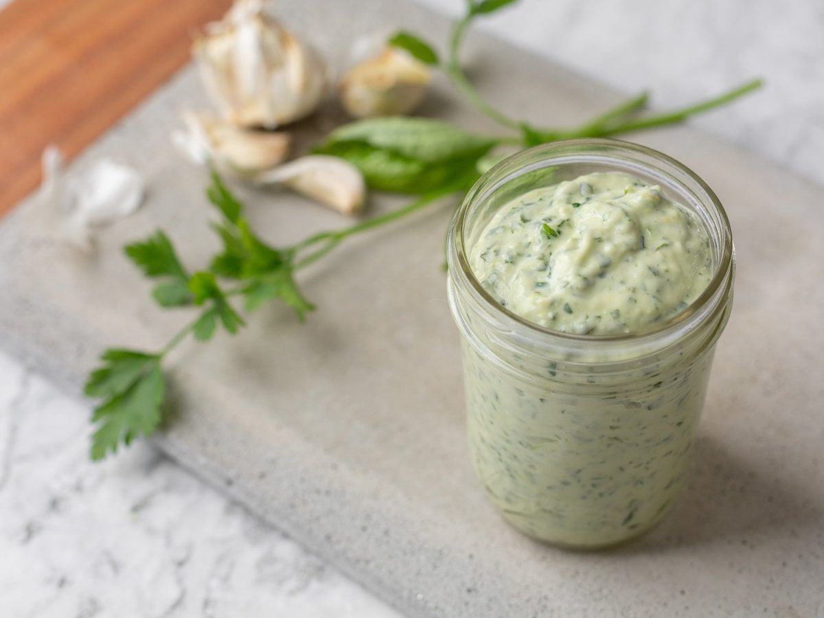 Ein Glas vegane Frankfurter Grüne Soße auf einem Schneidebrett, dahinter Kräuter und Knoblauch.