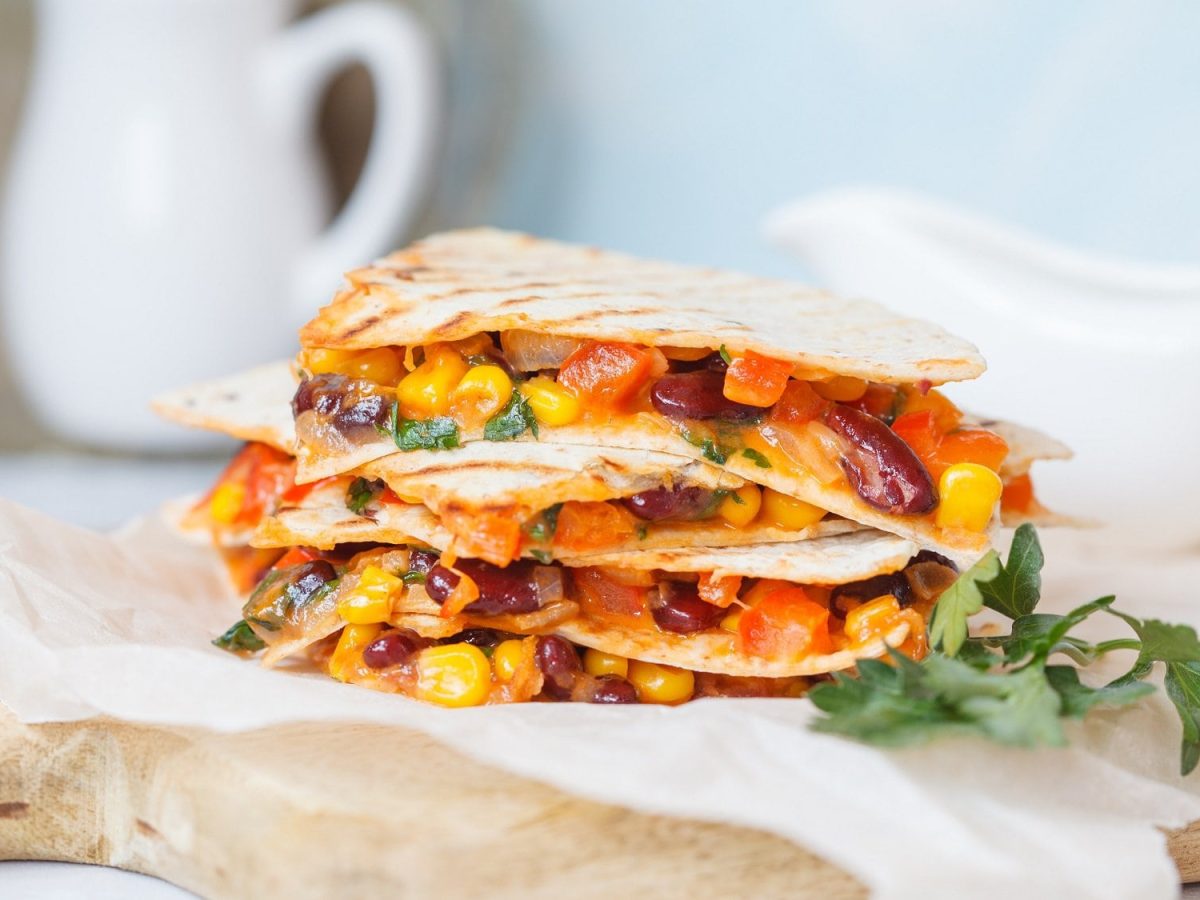 Ein Stapel vegane Quesadillas mit Mais, Paprika, Bohnen und mehr, auf Butterbrotpapier auf einem Brett.