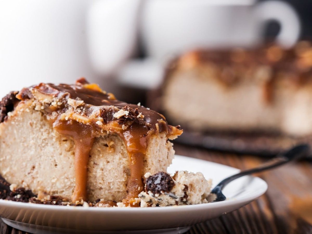 Ein Stück Erdnussbutter-Cheesecake mit Schokosauce und gehackten Erdnüssen garniert auf einem Teller. Im Hintergrund unscharf der restliche Kuchen.