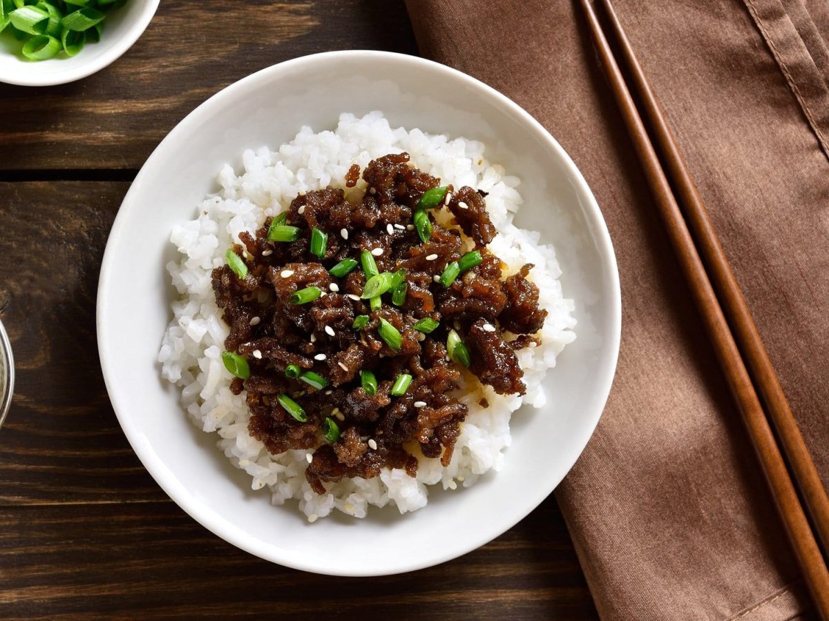 Ein Teller Reis und veganes Bulgogi mit Frühlingszwiebeln und Sesam bestreut in der Draufsicht, daneben Essstäbchen und Schalen mit Sesam und Frühlingszwiebeln.