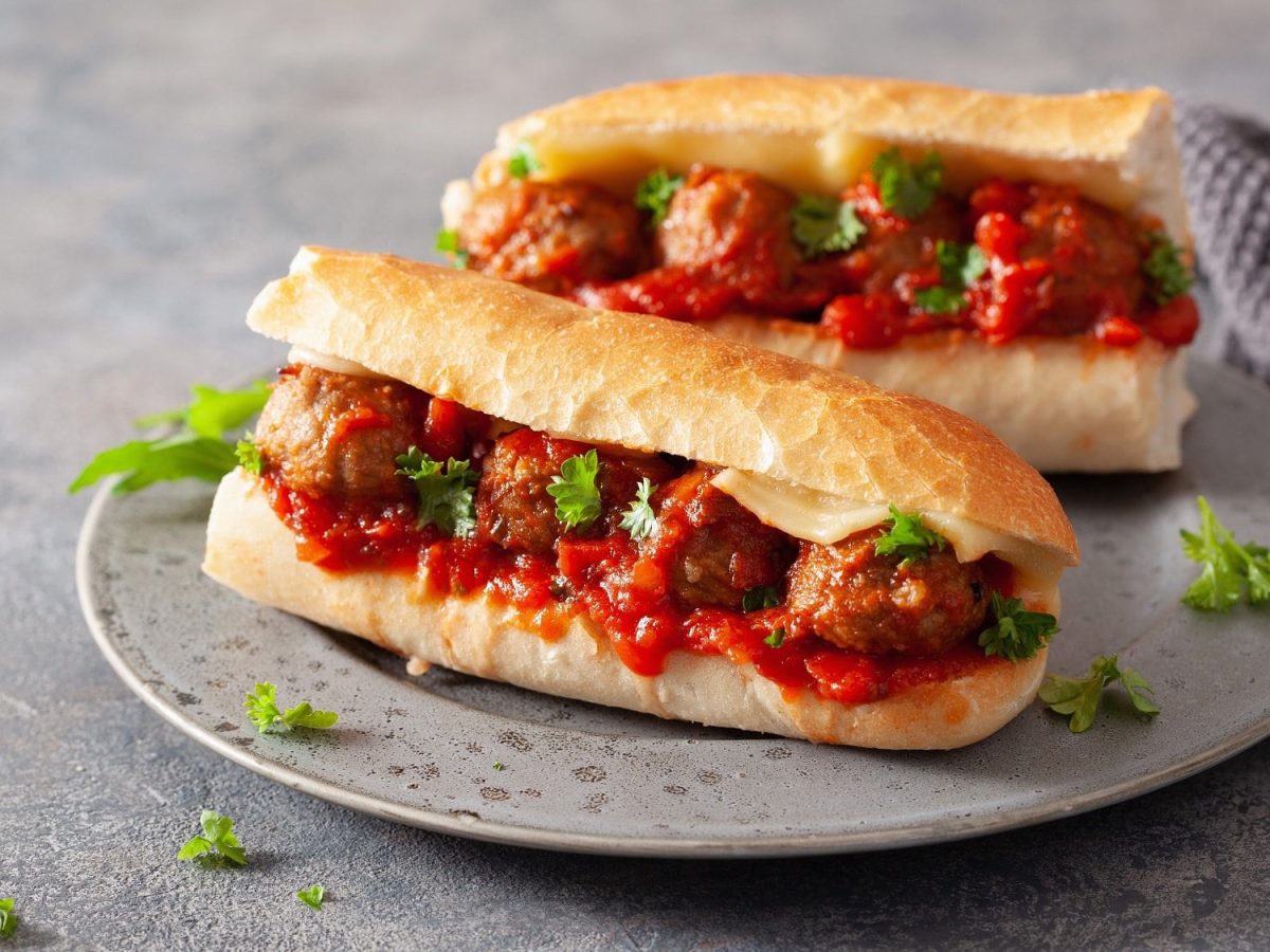 Zwei vegane Meatball Subs mit Tomatensauce, veganem Käse und Petersilie auf einem Teller.