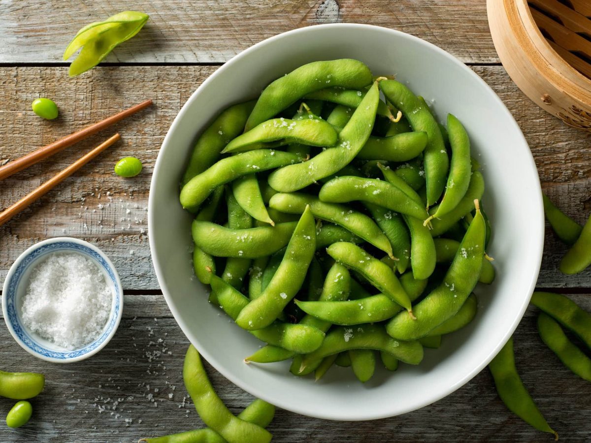 Draufsicht: Frisch gedämpfte Edamame mit Meersalz auf einer rustikalen Tischplatte bestreut.