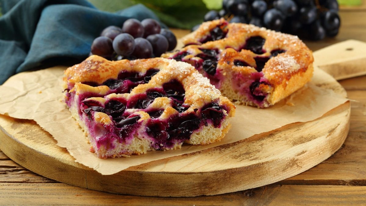 Zwei Stück Focaccia mit Weintrauben auf einem Holzbrett und einem Holztisch. Im Hintergrund zwei Bund dunkle Weintrauben.