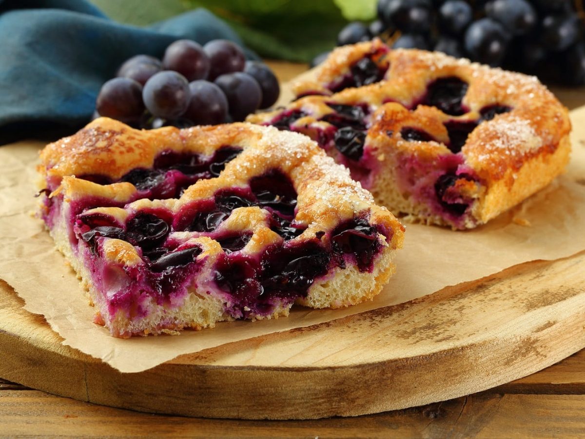 Zwei Stück Focaccia mit Weintrauben auf einem Holzbrett und einem Holztisch. Im Hintergrund zwei Bund dunkle Weintrauben.