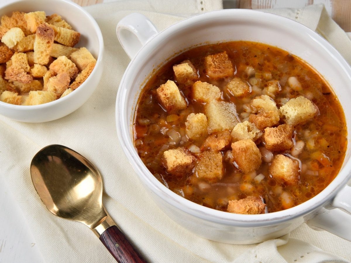 Draufsicht: Auf einem weißen Holztisch liegt ein weißes Tischtuch. Aus dem steht eine weiße Suppentasse mit einer Weiße-Bohnen-Kartoffel-Suppe. Daneben steht eine schale mit Croutons und daneben liegt ein Suppenlöffel.