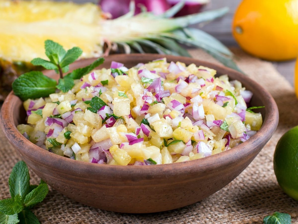 Eine braune Schale mit der Ananas-Salsa auf eionem Leinentuch mit Früchten im Hintergrund.