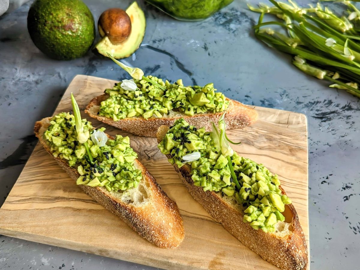 Bagutte mit dem Bärlauch-Avocado-Aufstrich auf einem Holzbrett mit dunkelgrauem Untergrund. Im Hintergrund Avocado und Bärlauch.