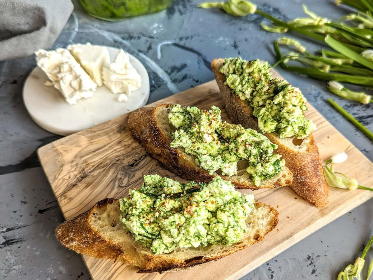 Ein kleines Holzbrett mit Baguettescheiben und der Bärlauch-Feta-Creme auf einem grauen Tisch mit Feta und Bärlauch im Hintergrund.