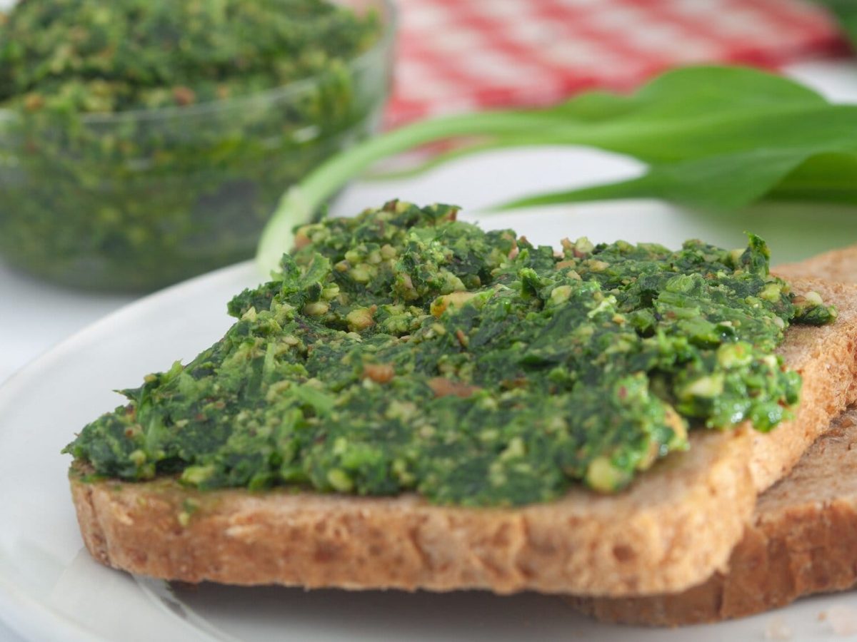 Frontalsicht: Ein Toast mit Bärlauch-Sardellen-Aufstrich liegt auf einem Holzbrettchen. Dahinter liegen Bärlauchblätter, sowie ein rot-weiß-kariertes Tuch.