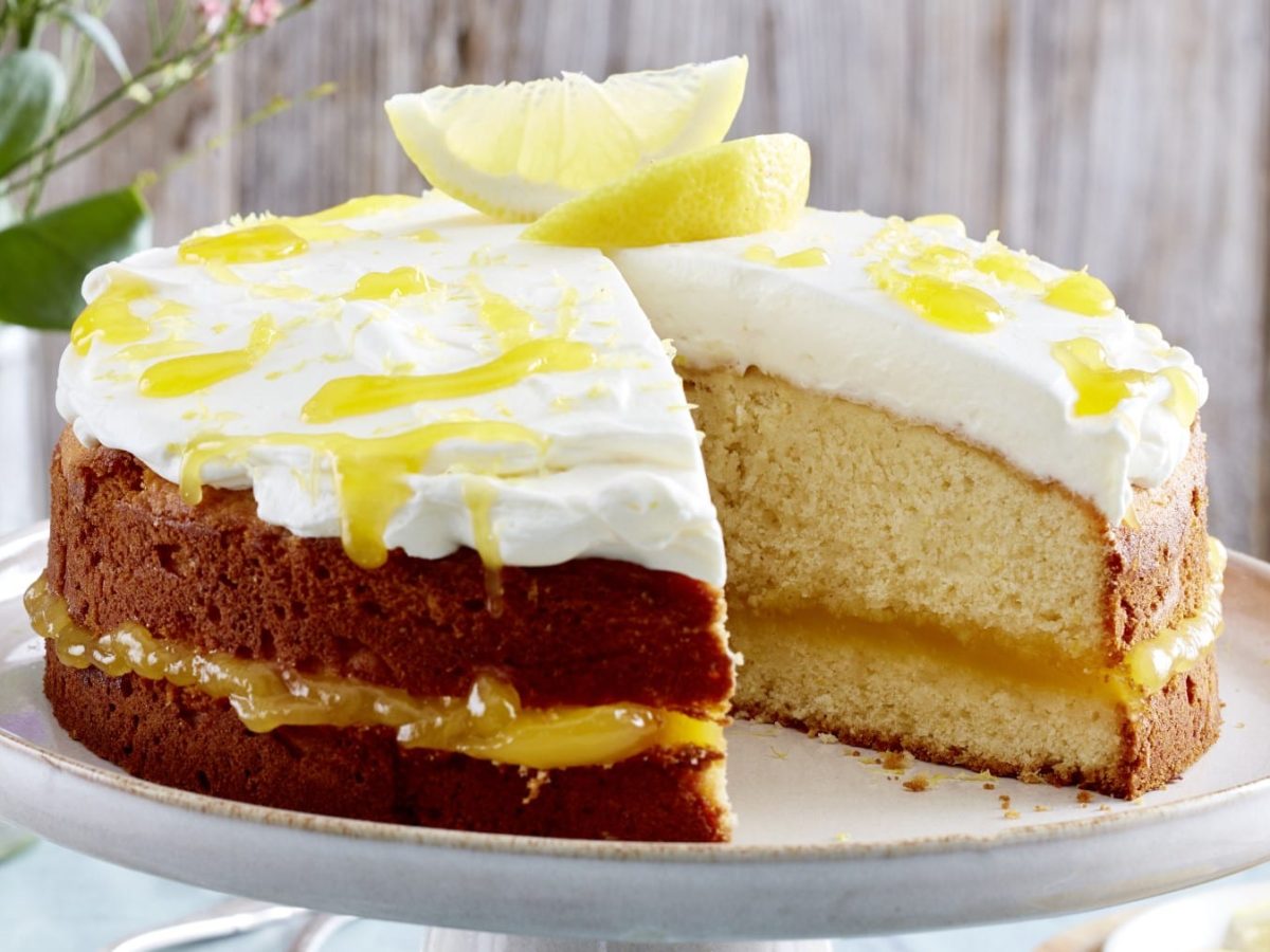 Angeschnittener Lemon-Curd-Kuchen mit Sahne und Zitronenspalten auf einem Servierteller, Holzwand und blaue Blumen im Hintergrund.