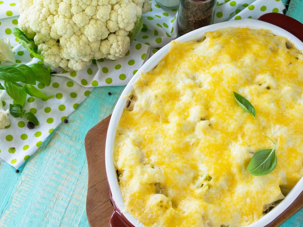 Draufsicht: Auf einem blauen Holztisch liegt ein Brettchen. Auf dem liegt eine ovale Auflaufform mit einem Blumenkohl-Lachs-Gratin mit Appenzeller. Neben der Form ist frischer Blumenkohl sowie eine Gabel.