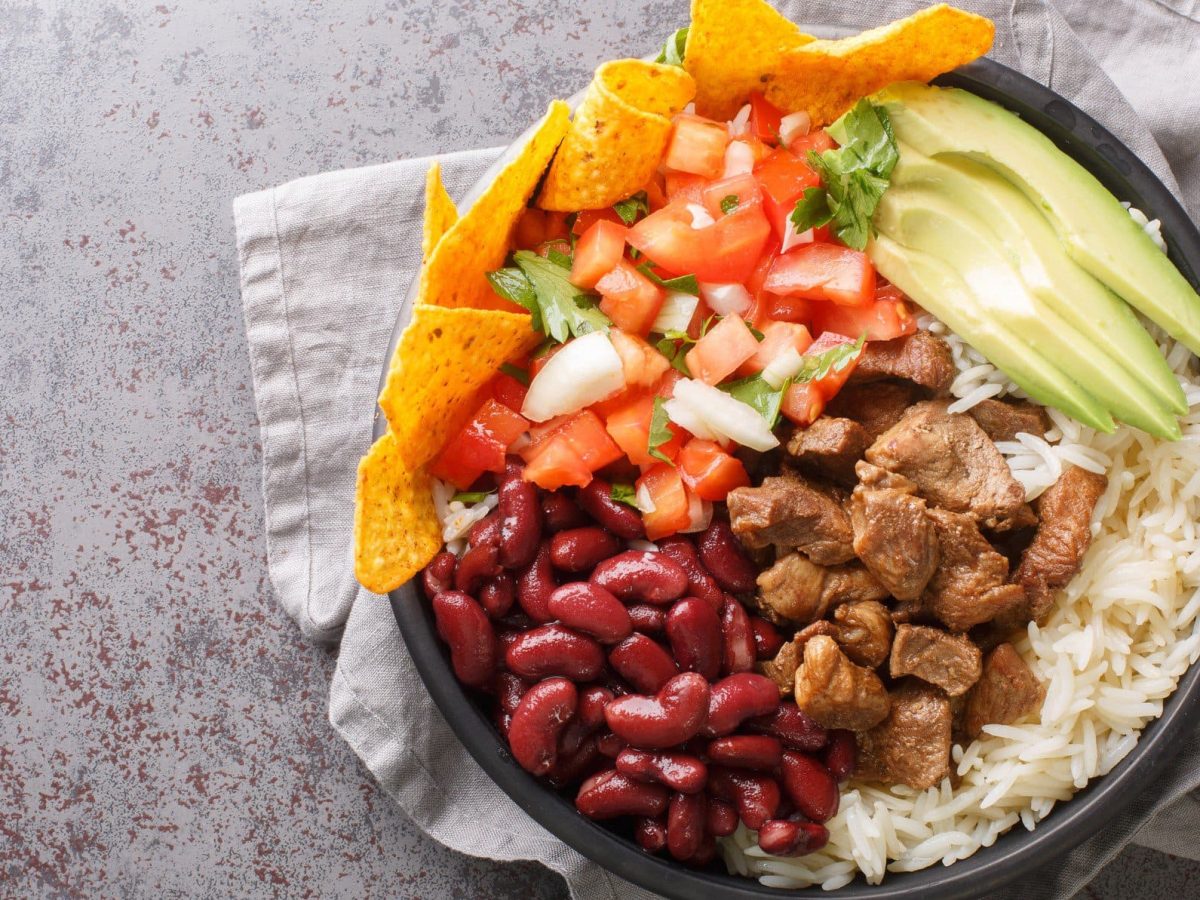 Chifrijo mit Chicharrón, Avocado, Tortilla-Chips, Bohnen und Reis in einer Schüssel auf grauem Tuch und Untergrund. Draufsicht.