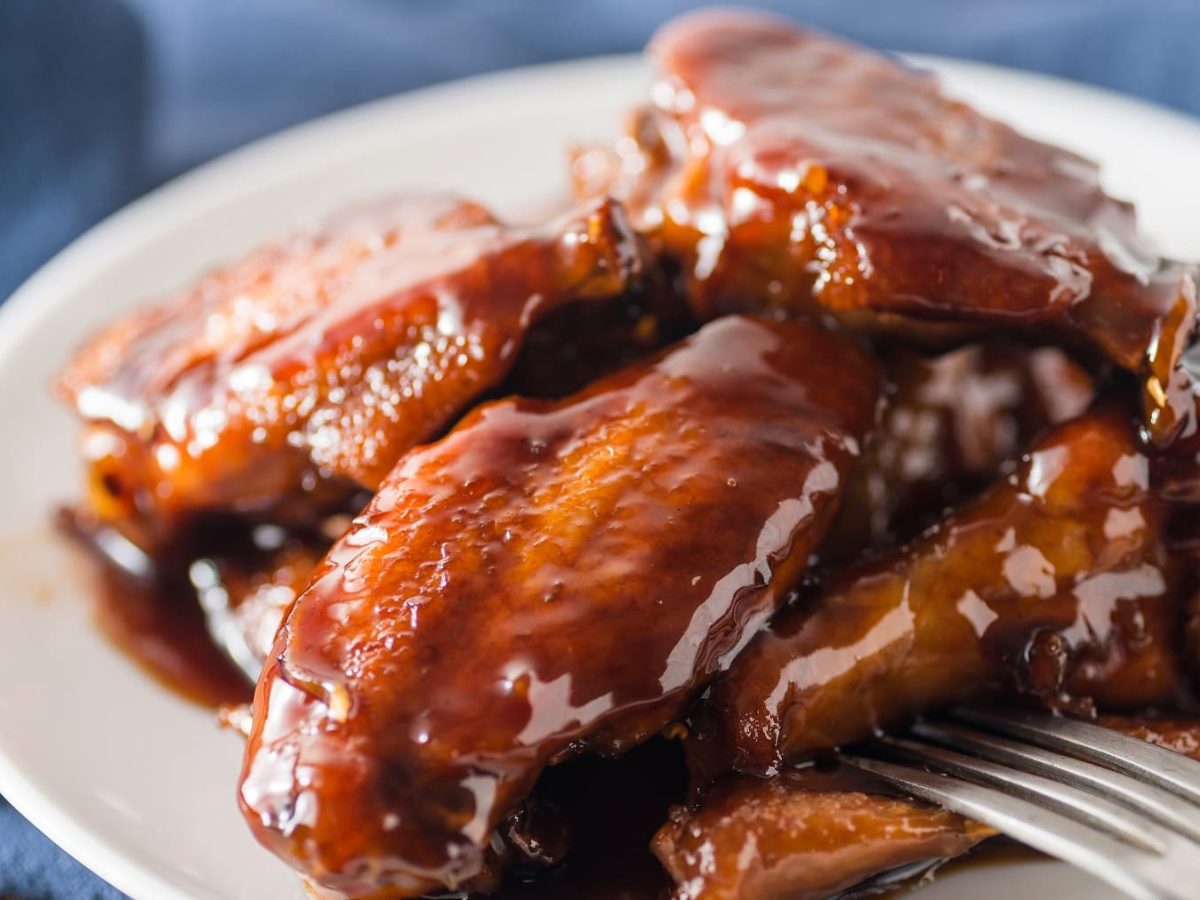 Ein weißer Teller mit den Cola-Chicken-Wings auf einem dunklen Holztisch mit einem blauen Tuch.