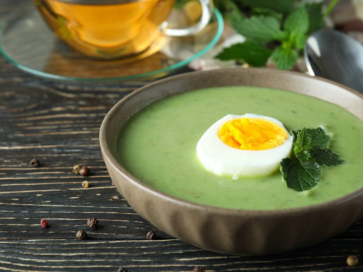 Draufsicht, schräg: Auf einem dunklen Untergund stehe eine Schüssel Brennnessel-Suppe mit Ei. Dahinter ist eine Tasse aus Glas mit einer goldigen Flüssigkeit drin.