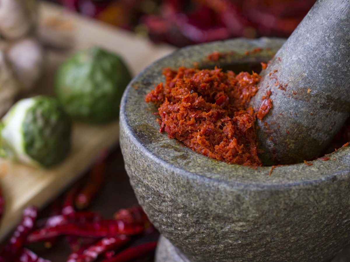 Ein Steinmörser mit Chilischoten, Limetten und Knoblauch unscharf im Hintergrund. Im Steinmörser eine rote Currypaste.