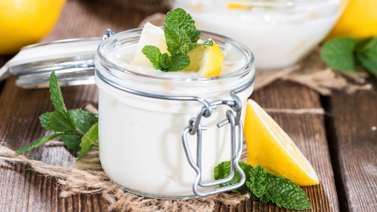 Frontal: Auf einem Holztisch steht ein Glas Zitronen-Buttermilch-Creme, die mit einer Zitrone und Minze garniert ist. Das Glas hat einen Deckel, der aber nicht aufliegt.