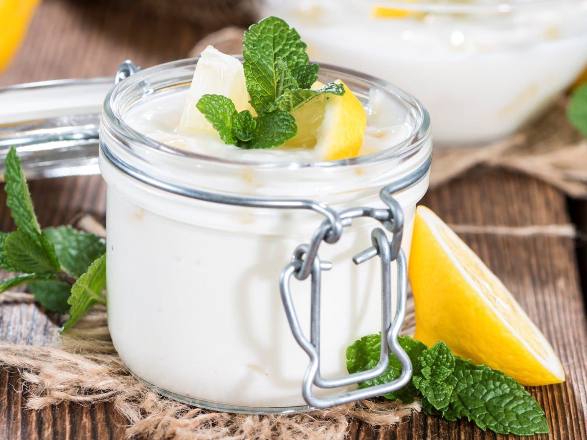 Frontal: Auf einem Holztisch steht ein Glas Zitronen-Buttermilch-Creme, die mit einer Zitrone und Minze garniert ist. Das Glas hat einen Deckel, der aber nicht aufliegt.