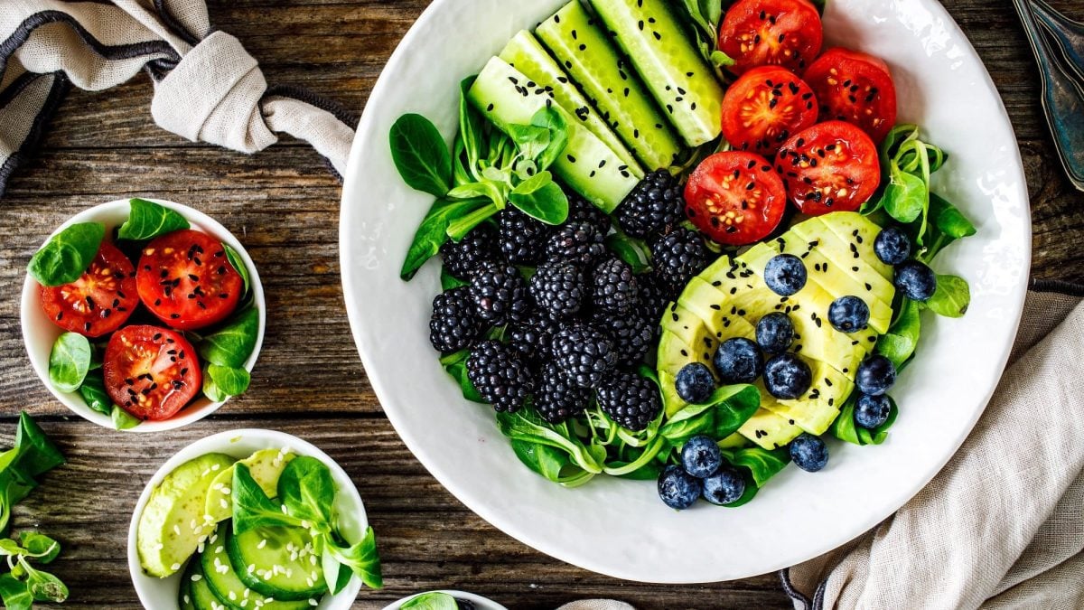 Draufsicht: Ein weißer tiefer Teller mit Feldsalat mit Avocado mit Beeren, Tomaten, Gurken, und schwarzem Sesam steht auf einem dunklen Untergrund aus Holz.