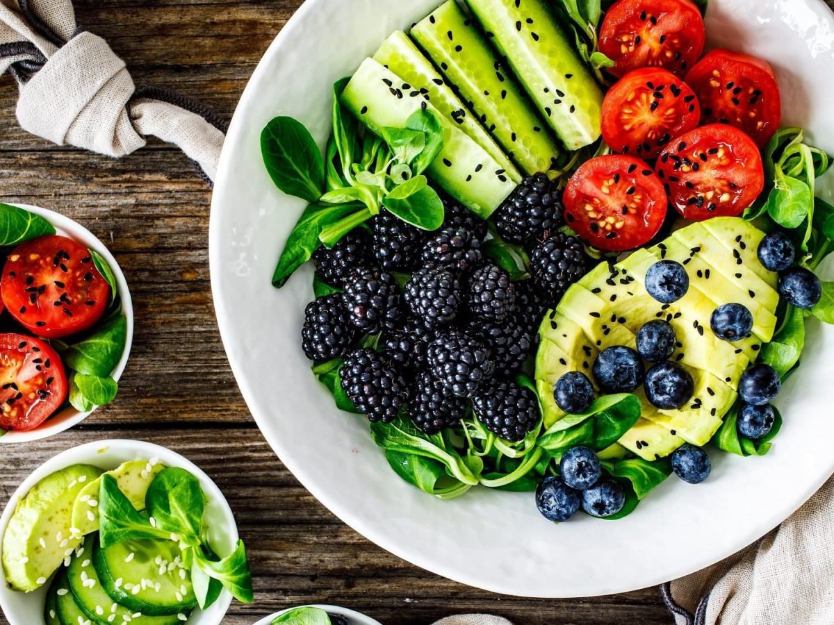 Draufsicht: Ein weißer tiefer Teller mit Feldsalat mit Avocado mit Beeren, Tomaten, Gurken, und schwarzem Sesam steht auf einem dunklen Untergrund aus Holz.