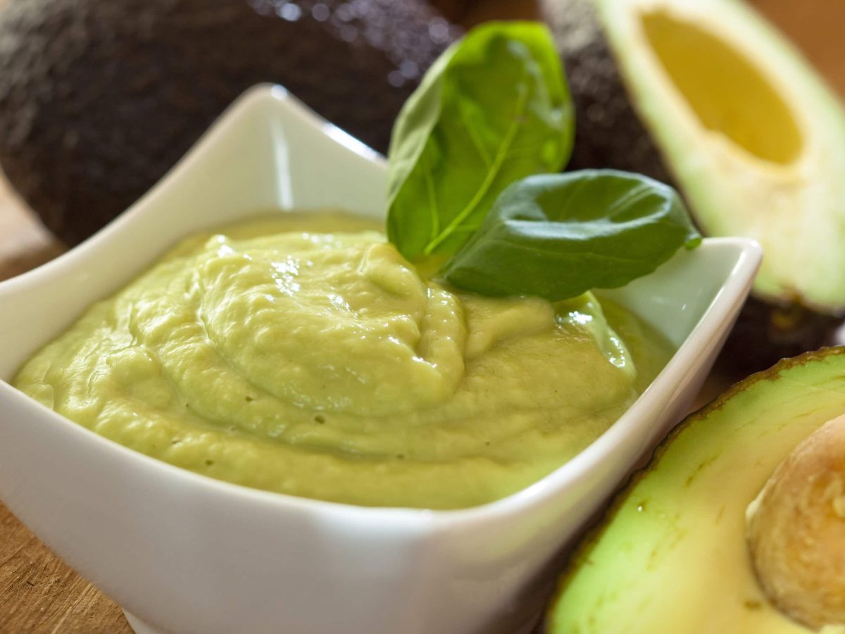 Feta-Joghurt-Creme mit Basilikum und Avocado in weißer Schale auf Holz. Im Bild Avocados und Basilikum. Draufsicht.