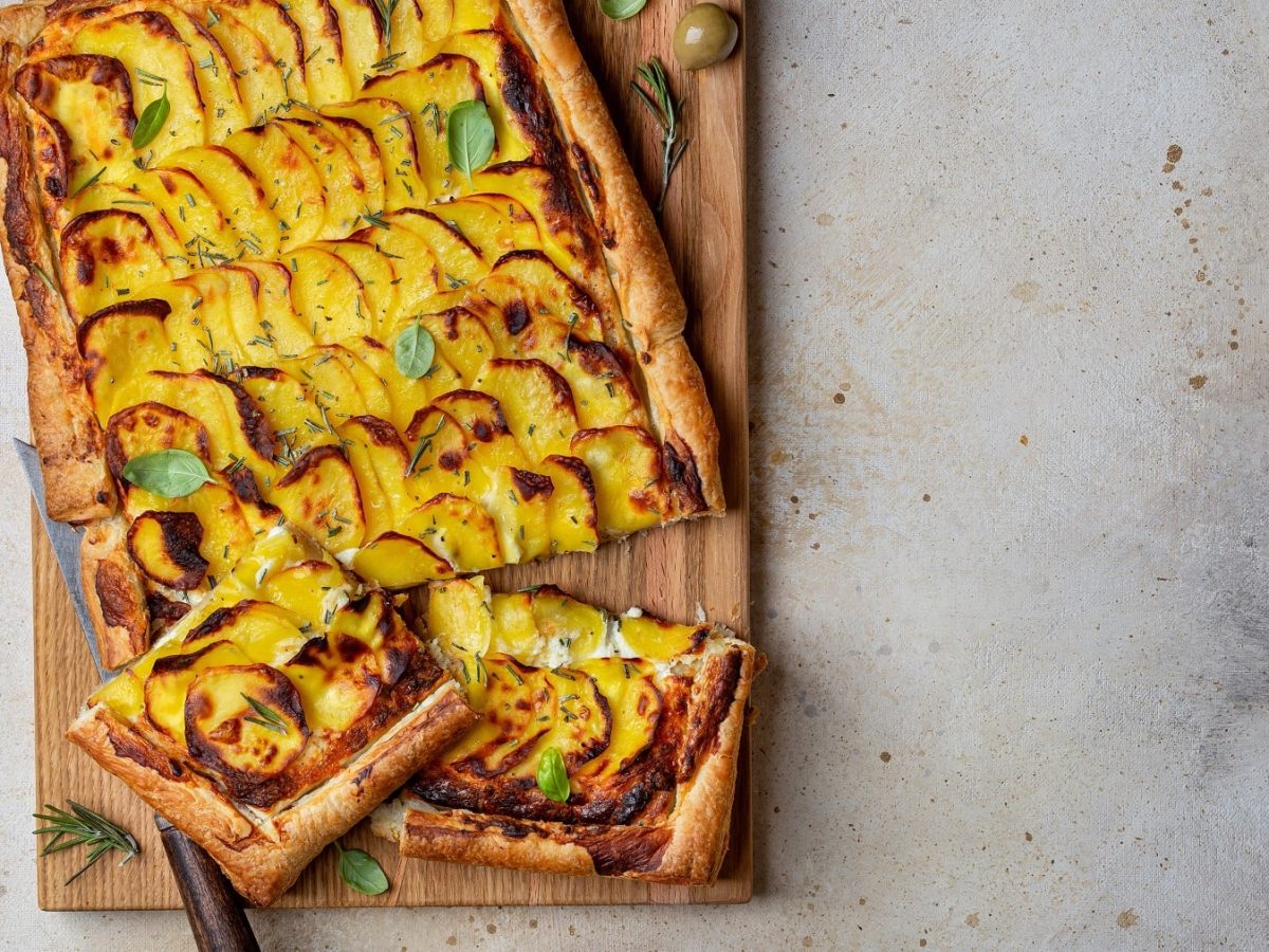 Ein Holzbrett mit dem Flammkuchen, der mit Kartoffeln und Rosmarin belegt ist auf hellem Steinuntergrund.